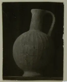 Pot with a long neck, single handle and vertical ribbing on the body, photographed against a dark background.