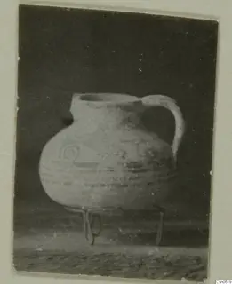 Round-bodied jug with a single handle and painted spiral and band decoration, standing on a three-legged wire stand.