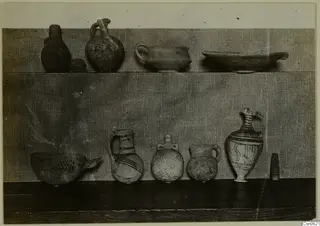 Several pottery vessels, some painted, arranged on two shelves against a cloth backdrop.
