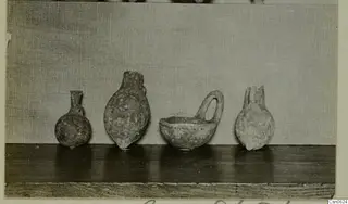 Four pottery vessels standing in a row on a wooden shelf, including a bowl with a loop handle.