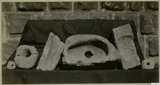 Group of worked stone objects on a dark cloth against a stone-block wall, including ring-shaped pieces and a basin-like stone.