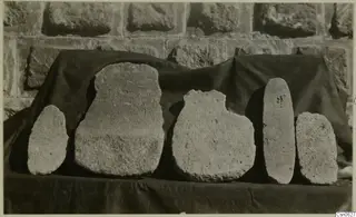 Five rough stone objects of varying shapes lined up on a dark cloth against a stone wall.