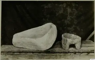 Two stone vessels on a wooden shelf: a large shallow trough and a small bowl standing on short feet