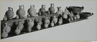 Many pottery jugs, juglets and two large bowls arranged in rows on wooden display shelves