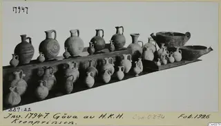 Pottery jars, juglets and two bowls displayed on wooden shelves against a pale background.