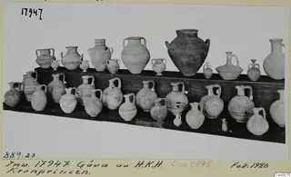 Two shelves of pottery jugs, jars and small juglets, some with painted bands, photographed on a photo-mount card