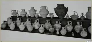 Group of pottery jars and juglets arranged in two rows on wooden shelves, many with handles and painted bands