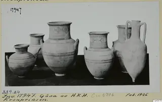 Six painted pottery jars and jugs standing in a row on a dark surface, photographed against a pale background.