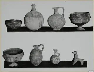 Two rows of painted pottery on dark shelves: jugs, footed bowls, a small jug and a small animal figurine