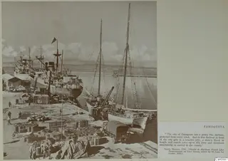 Harbour quay at Famagusta with moored ships, stacked barrels and crates, and a covered lorry beside two sailing vessels