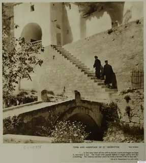 Stone steps up to a whitewashed building with an arched doorway, two robed men climbing, at the hermitage of St. Neophytos