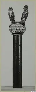 Tall dark cylindrical shaft topped by a head with an overlapping scale pattern and two small perched birds
