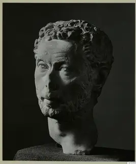 Carved stone male head with curly hair and a short beard, photographed against a dark background.