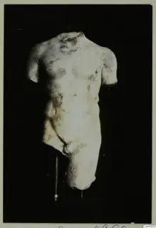 Pale stone torso of a male statue, head and arms missing, shown on a thin stand against a black background