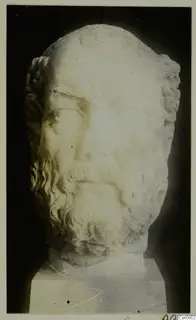 Marble head of a bearded man with curled hair, on a rectangular base against a dark background.