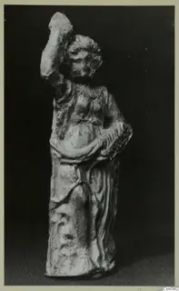 Standing statue of a figure in a long draped garment, one arm raised high, photographed against a dark background
