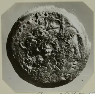 Find from Kountoura Trachonia: a corroded round metal object with raised markings, photographed face-on on a mounted card