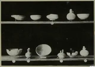 Two wooden shelves holding pale bowls and jugs against a black background, with round number labels