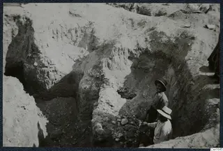 Excavation at Tombs 60-61, Marion, with two men in hats working among stones in a trench