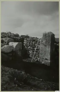 Dry-stone walls at Nitovikla with a tall upright stone block and a white measuring pole leaning against the wall