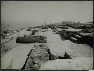 Excavated stone walls and paved floors at Vouni, with two people beside a ranging pole and the sea beyond