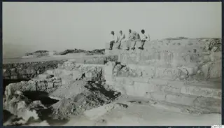 Five people sit in a row on top of a stone structure at Vouni, with cut stone courses and excavated ground below.