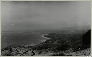 Coastal view from Vouni looking down over a curving bay, with low scrubby hills in the foreground and distant hills under a hazy sky.