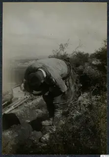 Man stooping over a dug cutting on a bushy slope at Marion, with an open landscape behind