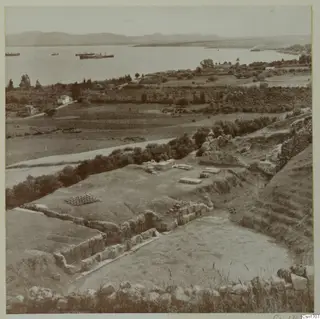 Excavated ruins at Soli with stone walls in the foreground, a bay with ships and hills beyond.