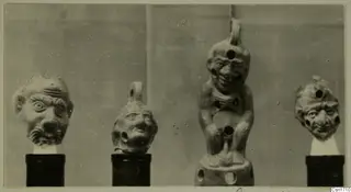 Four small terracotta figures and heads with exaggerated faces, each set on a stand against a grey backdrop.
