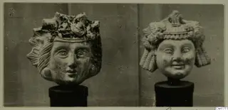 Two carved heads with elaborate headdresses, each set on a black cylindrical stand.