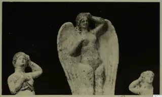 Three small pale figurines of women with arms raised to their heads, the central one with large spread wings, against a dark background