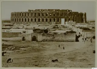 Large ruined structure with rows of arched openings rising above low houses, with people and animals on open ground in North Africa.