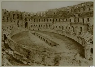 Ruined stone amphitheatre interior in North Africa, with tiers of arched openings around an oval arena.