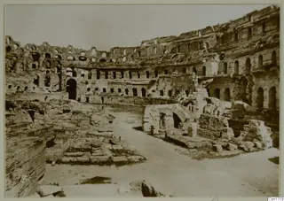 Interior of a ruined oval arena with tiers of arched openings and a stone vaulted structure on the right