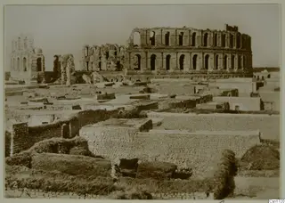 Ruined multi-storey arcaded structure rising above low walls of flat-roofed buildings, North Africa