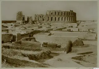 Ruined arched multi-storey structure rising above low walls and flat-roofed buildings