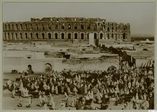 Large crowd of people and animals in an open space before a ruined multi-storey arcaded building, North Africa