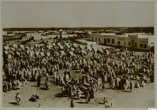 Crowded open square with many light tents and people in light robes, beside a long whitewashed building in North Africa