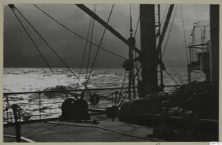 Ship's deck with ropes, winches and a railing, looking out over rough sea under a dark sky.