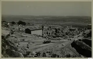 Wide view of a ruined site with standing columns, a shed, rail tracks with wagons and workers in the foreground, and hills beyond.