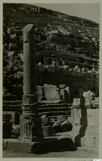 Tall stone column on a carved base beside stepped stone ruins, with a rocky hillside rising behind