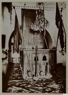 Interior of a building with a carved wooden enclosure, a hanging chandelier and flags on poles