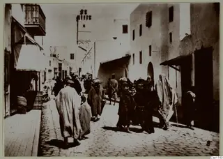 Street in North Africa with people in long robes, white buildings, an awning and a wooden balcony.
