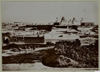 Town view of flat-roofed houses and domes, with camels and people on a track in the foreground.