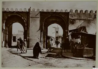 Street in North Africa passing through a crenellated gateway with arched openings, with pedestrians, a donkey with rider and a market stall