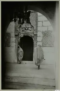 Arched doorway with patterned tilework, with two people in pale robes walking across the paved ground, North Africa