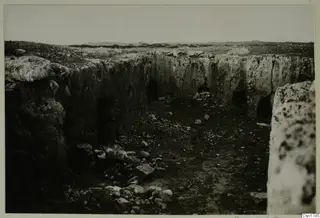 Rock-cut walls with dark openings and recesses in an excavated area, with rubble on the ground in front