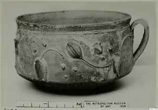Bowl with a loop handle and raised bud-like relief motifs and dots on its wall, photographed on a plain background with a scale bar.