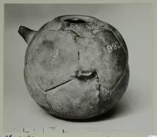 Globular vessel with a short spout on the left and a wide-rimmed opening at the top, photographed on a grey backdrop.